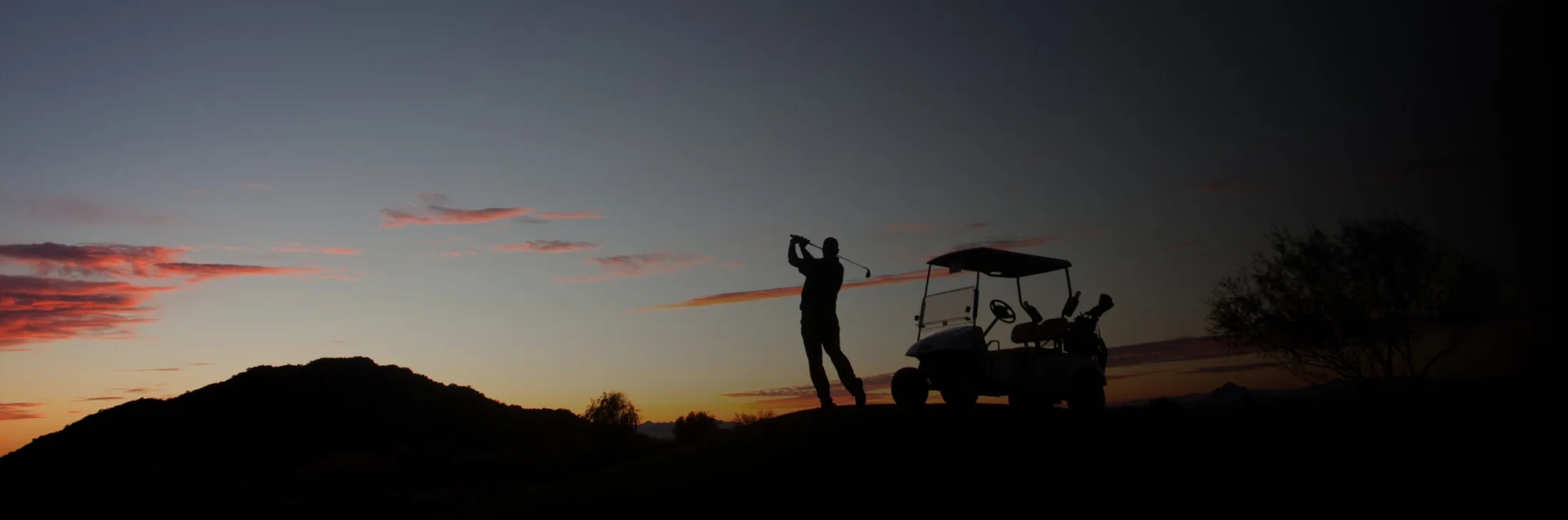 A golfer teeing off at dusk beside a cart